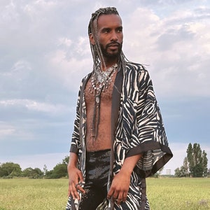 Könnte beinhalten: Eine Person trägt einen schwarz-weißen Zebra-Kimono mit einer silbernen Halskette und einem schwarzen Paillettenrock. Die Person steht auf einem Feld mit grünem Gras und einem bewölkten Himmel im Hintergrund.