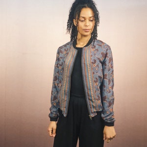 May include: A blue bomber jacket with a floral pattern in brown and orange. The jacket has a black collar, cuffs, and hem, and a zipper closure. The model is wearing a black top and pants.