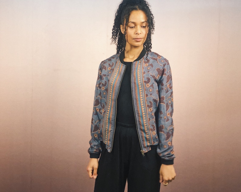 May include: A blue bomber jacket with a floral pattern in brown and orange. The jacket has a black collar, cuffs, and hem, and a zipper closure. The model is wearing a black top and pants.
