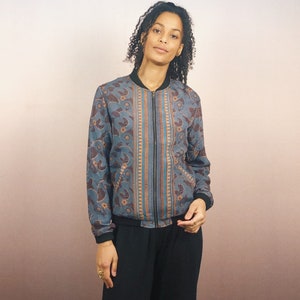 May include: A blue bomber jacket with a floral and paisley pattern in shades of brown and orange. The jacket has a black zipper, collar, and cuffs. The jacket is worn by a person.