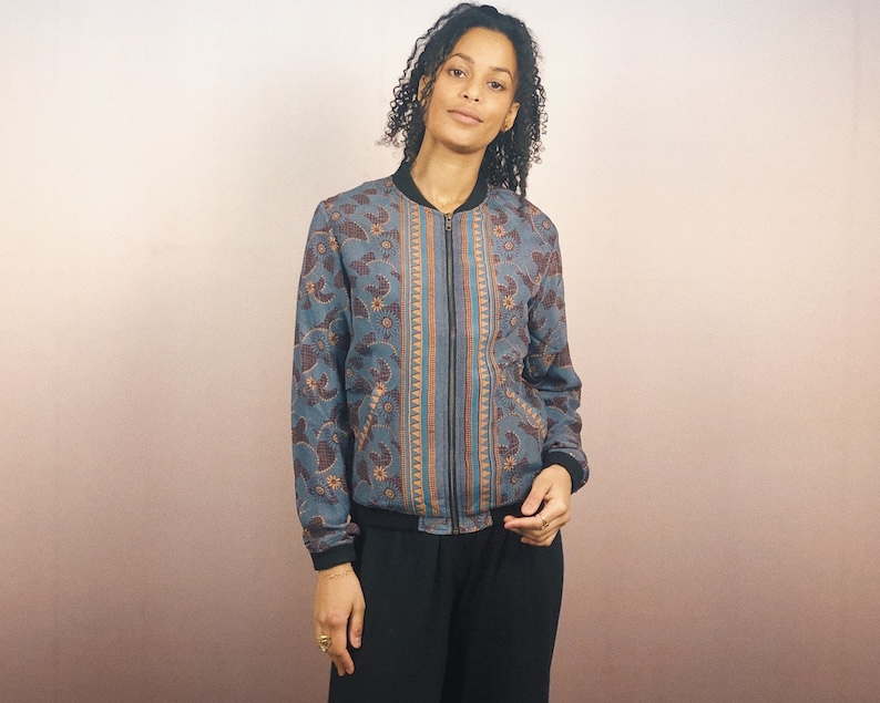 May include: A blue bomber jacket with a floral and paisley pattern in shades of brown and orange. The jacket has a black zipper, collar, and cuffs. The jacket is worn by a person.