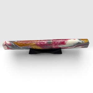 May include: A fountain pen featuring a marbled resin body with swirling patterns of magenta, white, grey, and metallic gold. The pen is cylindrical with a slightly tapered end and rests horizontally on a small, black, curved plastic display stand.