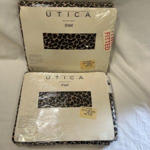 Puede incluir: Dos paquetes de sábanas de la marca Utica. Las sábanas tienen un estampado de leopardo en tonos marrones y negros. El embalaje incluye la marca, el tipo de sábana y la información de tamaño. Un paquete está etiquetado como "FITTED".