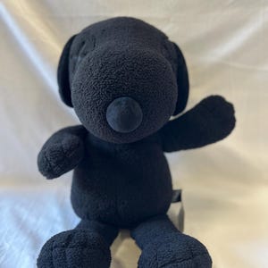 Op de afbeelding: Een zwart pluche hondenspeelgoed met slappe oren, een ronde neus en uitgestrekte armen. Het knuffeldier is gemaakt van een zacht, getextureerd materiaal en zit rechtop. Het speelgoed heeft een effen zwarte kleur.