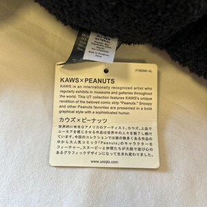 Op de afbeelding: Een wit label met zwarte tekst, met de KAWS x Peanuts-samenwerking. Het label beschrijft de kunstenaar KAWS en de Peanuts-personages in een gedurfde grafische stijl. De website www.uniqlo.com is ook opgenomen.