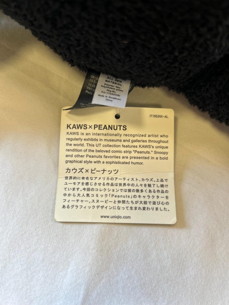 Op de afbeelding: Een wit label met zwarte tekst, met de KAWS x Peanuts-samenwerking. Het label beschrijft de kunstenaar KAWS en de Peanuts-personages in een gedurfde grafische stijl. De website www.uniqlo.com is ook opgenomen.