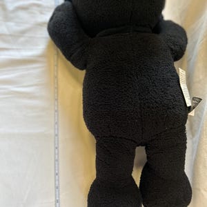 Op de afbeelding: Een zwarte teddybeer met een zachte, getextureerde buitenkant. De beer staat op een witte ondergrond, met een meetlint ernaast. De armen van de beer zijn omhoog en zijn benen zijn uitgestrekt.