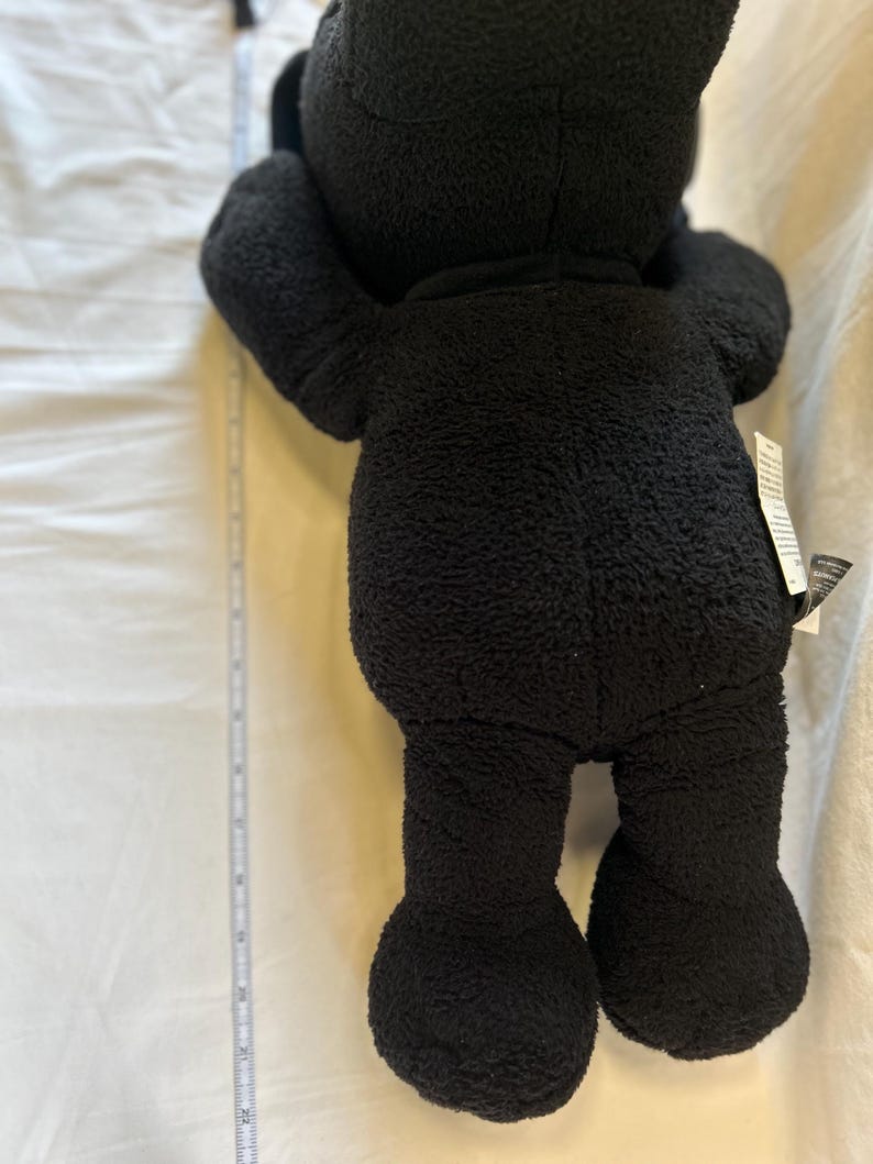 Op de afbeelding: Een zwarte teddybeer met een zachte, getextureerde buitenkant. De beer staat op een witte ondergrond, met een meetlint ernaast. De armen van de beer zijn omhoog en zijn benen zijn uitgestrekt.
