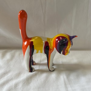 May include: A ceramic cat ornament with a raised tail. The cat is decorated with dripping paint in shades of orange, yellow, white, black, and purple. The cat is standing on four legs.