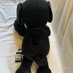 Op de afbeelding: Een zwarte pluche hond met grote oren en een rond lichaam. Het speelgoed heeft een label met de tekst "UNIQLO KAWS PEANUTS". De hond is gemaakt van een zacht, getextureerd materiaal.