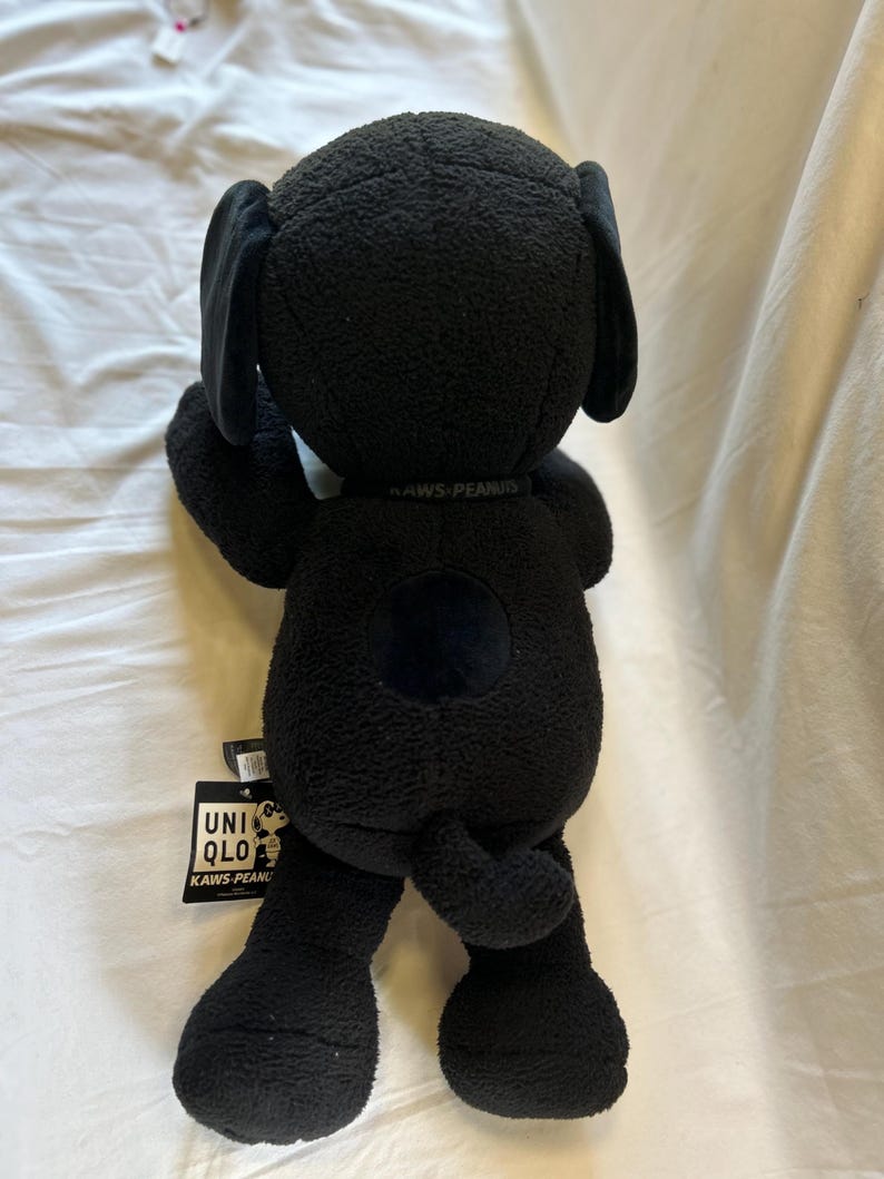 Op de afbeelding: Een zwarte pluche hond met grote oren en een rond lichaam. Het speelgoed heeft een label met de tekst "UNIQLO KAWS PEANUTS". De hond is gemaakt van een zacht, getextureerd materiaal.