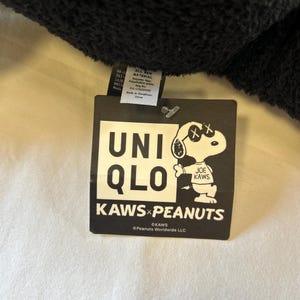 Op de afbeelding: Een zwart label met het Uniqlo-logo en de tekst "KAWS x PEANUTS". Het label toont een Snoopy-personage met doorgestreepte ogen, een handtekening van de kunstenaar KAWS. Het label bevat ook materiaalinformatie.