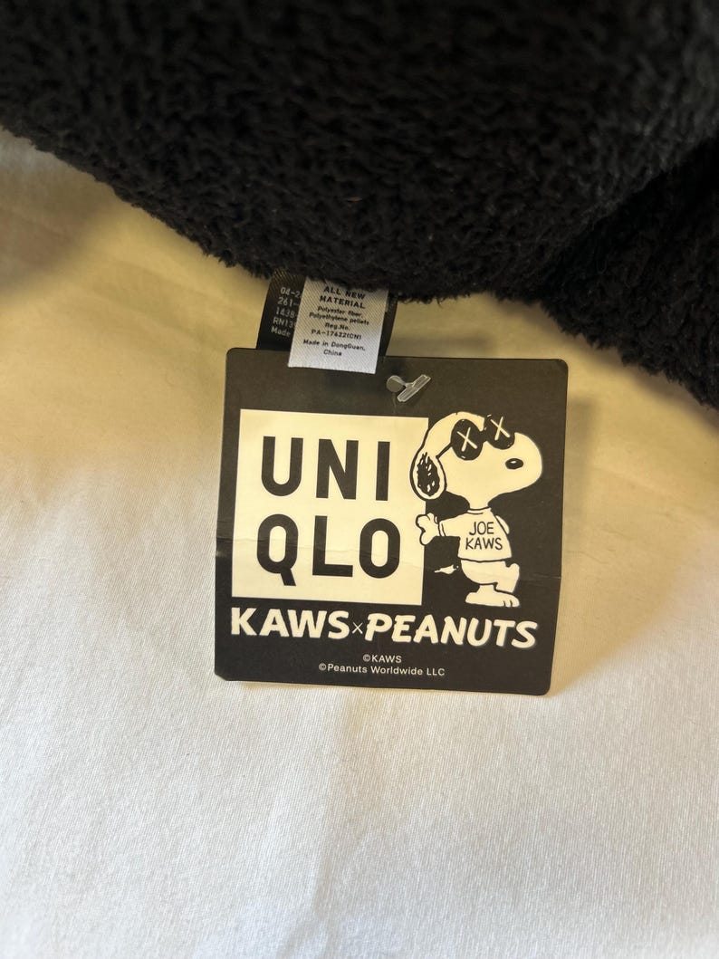 Op de afbeelding: Een zwart label met het Uniqlo-logo en de tekst "KAWS x PEANUTS". Het label toont een Snoopy-personage met doorgestreepte ogen, een handtekening van de kunstenaar KAWS. Het label bevat ook materiaalinformatie.
