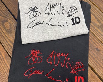 May include: Two t-shirts with the signatures of the members of the band One Direction. The signatures are embroidered in black on a gray t-shirt and in red on a black t-shirt. The signatures include Harry, Niall, Liam, and Zayn. The t-shirts are stacked on top of each other.