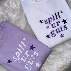 Puede incluir: Dos sudaderas con el texto "spill ur guts" bordado en morado en una sudadera blanca y una sudadera lavanda. Ambas sudaderas tienen estrellas moradas alrededor del texto.