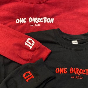 Może przedstawiać: Dwie bluzy, jedna czerwona, a druga czarna, z wyszytymi na przodzie napisami "ONE DIRECTION est. 2010". Czerwona bluza ma białe haft, a czarna ma czerwony haft. Obie bluzy mają wyszytą na rękawie cyfrę "1D".