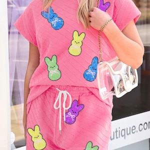 Può includere: Completo da salotto rosa a due pezzi con motivo strutturato e applicazioni di coniglietti colorati. Il set include un top a maniche corte e pantaloncini con vita con coulisse. È visibile anche una borsa trasparente con catena dorata.