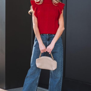 May include: A woman wearing a red turtleneck top with ruffle sleeves and blue jeans. She is holding a beige purse with a gold chain strap. The outfit is completed with beige shoes and sunglasses.