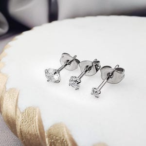 May include: Three silver-toned stud earrings featuring clear, round-cut stones in four-prong settings. Each earring includes a matching metal butterfly-style back. The earrings are arranged in a row, showing a graduated size from largest to smallest.