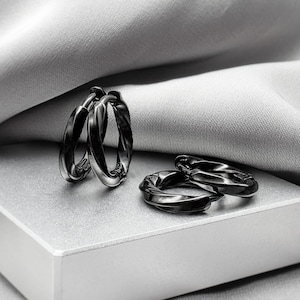 Puede incluir: Un conjunto de tres pares de pendientes de aro negros. Dos pares están planos, mientras que el tercer par está de pie. Los pendientes tienen un diseño retorcido y se muestran en una caja blanca con un fondo de tela gris.