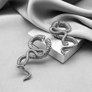 May include: Two silver-toned snake earrings. Each earring is a coiled snake with detailed scales, a small head, and a curved tail. The earrings are displayed on a white surface against a gray background with draped fabric.