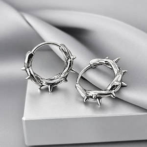 May include: A pair of silver-toned hoop earrings featuring a textured, vine-like design with multiple small, pointed spikes protruding from the outer surface. The metal has an oxidized finish that highlights the intricate, rugged details of the hoops.