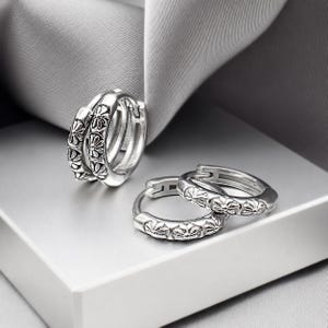 May include: A pair of silver hoop earrings featuring a repeating embossed floral or cross-like pattern along the outer surface. The earrings have a hinged closure mechanism and a polished metallic finish, showing intricate detailing on the metal.