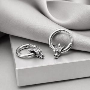 May include: A pair of silver-toned hoop earrings, each featuring a detailed dragon head charm. The earrings have a polished metallic finish and a circular hoop design. The dragon heads are sculpted with visible scales and facial features.