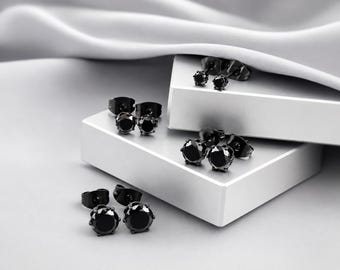 May include: A collection of three pairs of black stud earrings, each featuring a round, faceted stone set in a dark metal prong setting. The earrings are displayed in varying sizes, with two pairs resting on white blocks and one pair on the surface.
