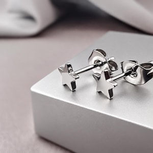 May include: A pair of silver-toned star-shaped stud earrings with a polished metallic finish. Each earring features a five-pointed star design attached to a straight post with a secure butterfly-style backing, reflecting light on the smooth surface.