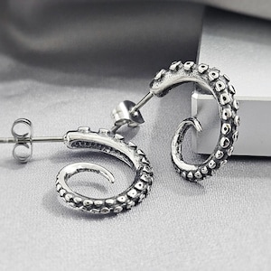 May include: A pair of silver-toned stud earrings shaped like curled octopus tentacles. Each earring features a textured surface with raised circular suction cups and a post back closure, finished with an oxidized effect to highlight the details.