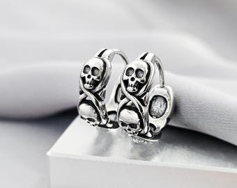 May include: A pair of silver hoop earrings featuring a detailed relief design of multiple skulls. The metal has an oxidized finish to highlight the intricate facial features of the skulls and the surrounding curved, rope-like patterns.