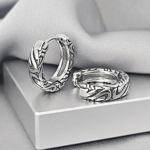 May include: A pair of silver hoop earrings featuring a raised, intricate, swirling relief pattern across the outer surface. The earrings have a hinged closure mechanism and a polished metallic finish with darkened recessed areas to highlight the design.