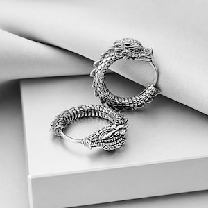 May include: A pair of silver-toned hoop earrings featuring a detailed dragon design. Each earring is shaped like a coiled dragon with textured scales covering the entire hoop, ending in a sculpted dragon head with visible eyes and snout details.