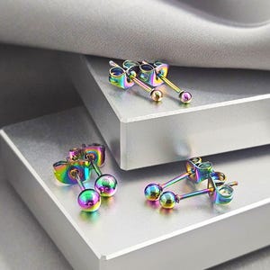 May include: Three pairs of stud earrings featuring a polished ball design. Each earring is crafted from metal with an iridescent rainbow finish that shifts between shades of blue, green, purple, and gold. The set includes matching butterfly backs.