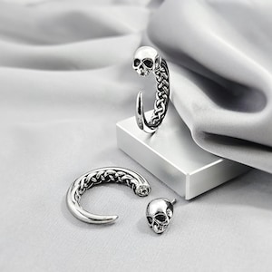 May include: Silver skull jewellery set. The set features a curved earring with a skull and braided design, a matching curved piece, and a separate skull charm. The pieces are displayed on a white surface against a grey background.