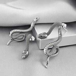 May include: A pair of silver-toned snake earrings featuring detailed scale textures and small, vibrant green inset eyes. The earrings are designed with a post-back closure, showcasing a coiled serpent shape that curves elegantly to create a distinct look.
