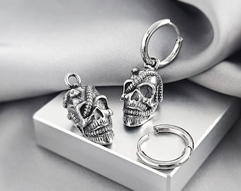 May include: Two silver-toned metal skull charms, one attached to a circular hoop earring, and one separate circular hoop earring. Each skull features a detailed, textured surface with a snake coiled around the cranium and eye sockets.