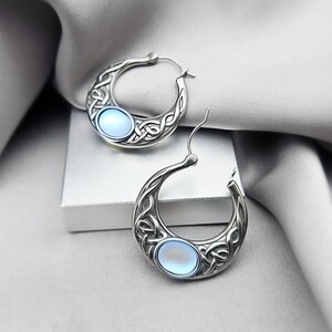 May include: A pair of silver-toned hoop earrings featuring an intricate interlaced knot pattern and a single oval-shaped, light blue cabochon stone set into the lower curve of each earring. The earrings have a polished metallic finish and curved posts.