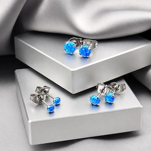 May include: Three pairs of stud earrings featuring round, vibrant blue opal-like stones set in polished silver-tone metal. Each earring has a secure post back and a prong setting that holds the circular stone firmly in place for a classic look.
