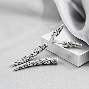 May include: Silver-toned stud earrings with a Celtic knot design. The earrings have a long, pointed shape with intricate detailing. One earring is detached, and the others are on a white surface, with a gray background.