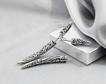 May include: A pair of silver-toned earrings featuring a curved, tapered spike design with intricate, raised knotwork patterns. The earrings have a polished metallic finish with darkened recessed areas that highlight the detailed, flowing engraved motifs.