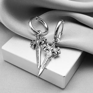 May include: A pair of silver-toned hoop earrings. Each earring features a dagger-shaped pendant with ornate, swirling details. The earrings are displayed on a white surface with a gray, draped fabric in the background.