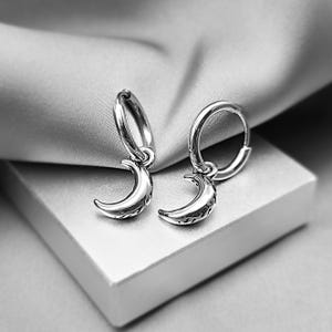 May include: A pair of silver-toned hoop earrings featuring dangling crescent moon charms. Each moon charm has a polished, reflective surface with intricate, dark-toned engraved detailing along the inner curve of the crescent shape.