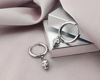 May include: A pair of silver-toned hoop earrings, each featuring a small, dangling skull charm. The earrings have a polished metallic finish and a hinged closure mechanism, presenting a clean, reflective surface on both the hoops and the skull pendants.