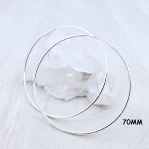 May include: A pair of thin, circular silver hoop earrings with a smooth, polished finish. The earrings have a diameter of 70mm as indicated by the text printed in the lower right corner. The hoops are positioned in front of a clear crystal.