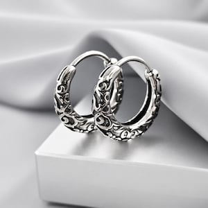 May include: A pair of silver hoop earrings featuring an intricate, raised scrollwork pattern across the outer surface. The metal has a polished finish with darkened recessed areas to highlight the detailed relief design of the circular hoops.