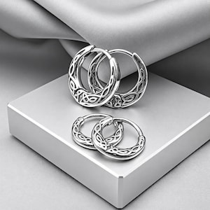 May include: Two pairs of silver hoop earrings featuring an intricate, raised Celtic knotwork pattern. The larger pair is positioned behind the smaller pair, both displaying a polished metallic finish with dark recessed detailing.