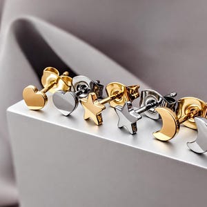 May include: A collection of six stud earrings featuring heart, star, and crescent moon shapes. The set includes three gold-toned and three silver-toned metal earrings, each equipped with a standard post and a secure butterfly clutch backing.