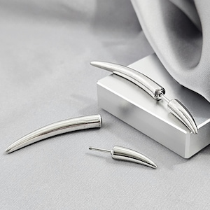 May include: A set of silver-toned earrings featuring a curved, tapered spike design. One earring is assembled with a post and back, while the other is shown as two separate pieces, including a long curved spike and a smaller matching spike component.